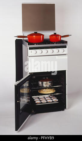 Gas cooker with two red pots on hob, oven door open to reveal uncooked cakes in trays on shelves. Stock Photo