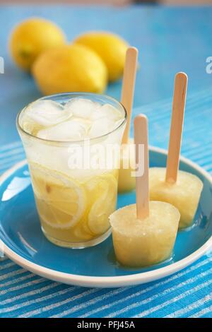Glass of lemonade and three lemonade ice lollies on a plate, with fresh lemons in the background, close-up Stock Photo