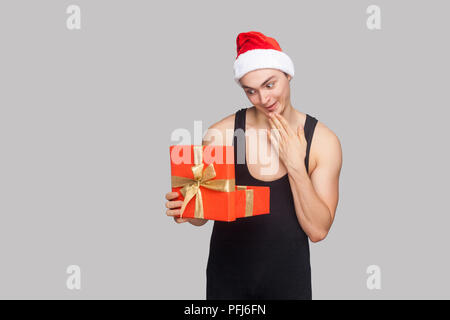 New year concept. Man in red hat holding red gift box and looking inside with amazed funny face. Indoor, studio shot, isolated on gray background Stock Photo