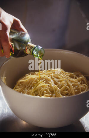 Olive oil being poured from bottle over cooked spaghetti in white bowl. Stock Photo