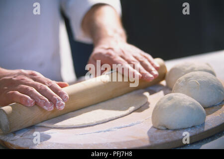 Three dough balls resting on large wooden board, thin layer of dough being rolled out with wooden rolling pin Stock Photo