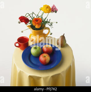 Small round table laid with red mug, colourful flowers in yellow jug, three apples on blue plate, pear,  elevated view. Stock Photo