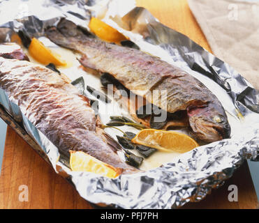 Two whole trouts cooked in foil, stuffed with sage leaves and garnished with lemons Stock Photo