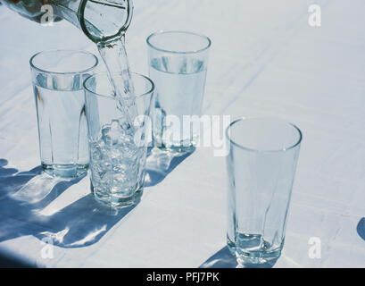 Four tall clear glasses standing on white table cloth, two filled with water, water being poured from glass jug into empty glass Stock Photo