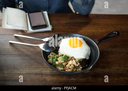 Woman with cell phone having stir fried chicken chilli and thai basil with fried egg on top in restaurant Stock Photo