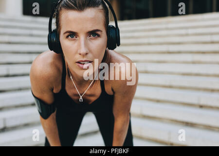 fitness woman training outdoors bending forward with hands on knees. fitness woman wearing wireless headphones relaxing while training. Stock Photo