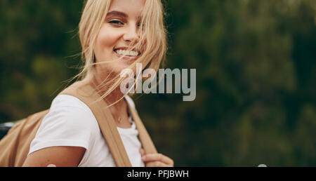 Close up of woman on a holiday walking in a park wearing a bag. Portrait of a smiling woman with brown hair flying over her face. Stock Photo