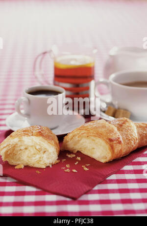 Croissant broken in half, cup of espresso, cup of coffee, glass of tea and jug of milk, laid out on red napkin and checked tablecloth, close-up Stock Photo