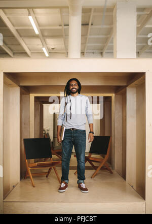 Smiling businessman holding laptop standing inside an enclosure with seats at office. Businessman with long hair standing with a laptop. Stock Photo