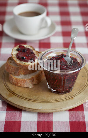 Home-made mixed berry jam in jar and on slice of white bread, served on wooden chopping board, next to cup of tea, checked tablecloth Stock Photo