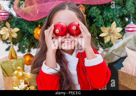 Close up of smiling girl wearing a red santa costume and holding two christmas balls in her hands and posing over her eyes, with a Christmas tree behind, christmas concept Stock Photo
