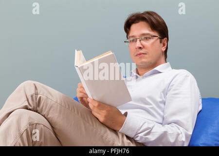 view of handsome young businessman in casual clothes and eyeglasses reading a book while lying on couch at home Stock Photo