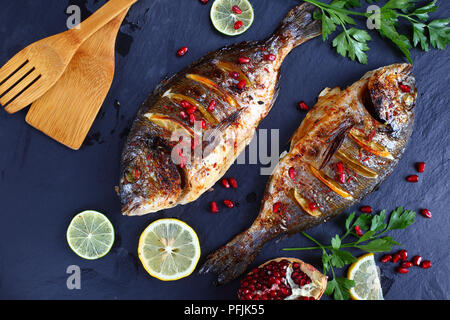 delicious roasted spicy Gilthead seabream fish with lemon slices and fresh parsley, pomegranate seeds on slate tray with wooden shovels,  horizontal v Stock Photo