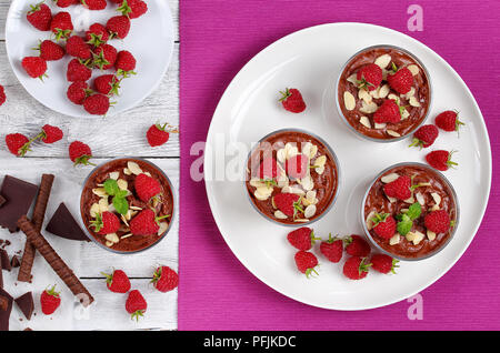 delicious chocolate mousse in glass cups topped with fresh mint leaves, raspberries and almond flakes on white plate on table mat with ingredients on  Stock Photo