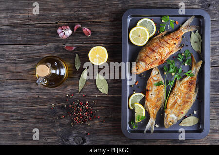 delicious roast  bream fish marinated with lemon slices, bay leaf, spices on baking sheet on old dark rustic table with ingredients on background, hor Stock Photo