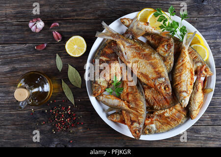 delicious hot fried mix of freshwater fish marinated with lemon slices, bay leaf, spices on white platter on old dark rustic table, horizontal view fr Stock Photo