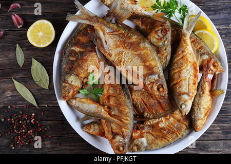 close-up of delicious hot fried mix of freshwater fish marinated with lemon slices, bay leaf, spices on white platter on old dark rustic table, horizo Stock Photo
