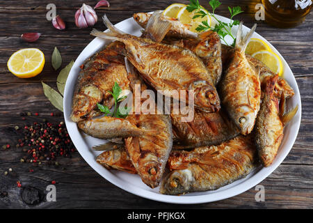 close-up of delicious roast mix of freshwater fish marinated with lemon slices, bay leaf, spices on white platter on old dark rustic table, horizontal Stock Photo