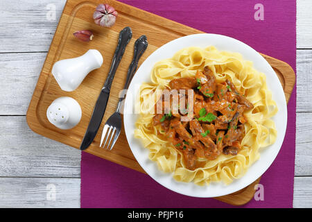 fettuccine pasta topped with delicious beef stroganoff stewed in sour cream sauce with porcini mushrooms on white plate on cutting board with cutlery, Stock Photo