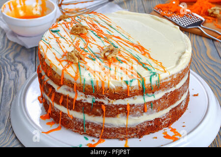 delicious carrot cake with cream cheese frosting decorated with walnuts and drizzled with colorful ganache on white platter on wooden table with ingre Stock Photo