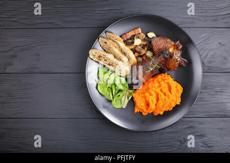 a portion of Coq Au Vin - classic French chicken stew served on black plate with mix of lettuce leaves and pumpkin puree, horizontal view from above Stock Photo