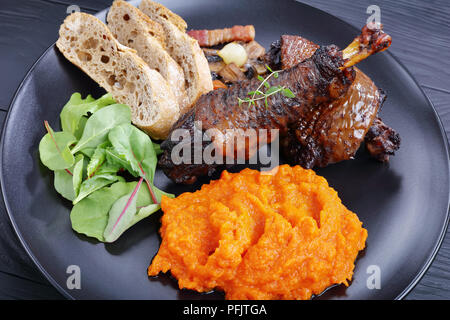 a portion of Coq Au Vin - classic French chicken stew served on black plate with mix of lettuce leaves, pumpkin puree and bread, horizontal view from  Stock Photo