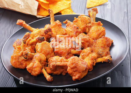 delicious deep fried battered crispy chicken winglets with exposed bones on a black plate on black wooden table. nachos in paper bag at background, vi Stock Photo