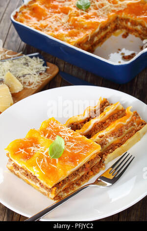 portion of italian lasagna topped with melted cheese and fresh basil leaves, sprinkled with grated parmesan cheese on white plate with fork, authentic Stock Photo