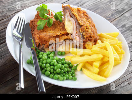 delicious Cordon Bleu with French fries, green peas and parsley on plate on old dark wooden table with silver fork and knife, authentic recipe, close- Stock Photo