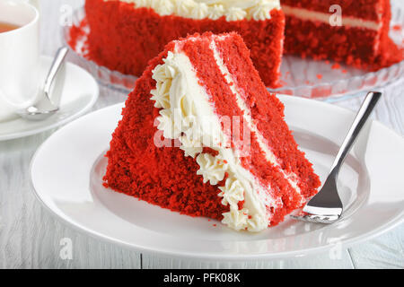 a slice of red velvet cake topped with beautiful creamy roses on plate on wooden table with cup of tea and whole cake at background,  view from above, Stock Photo