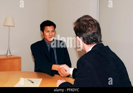Two businessmen shaking hands across desk Stock Photo