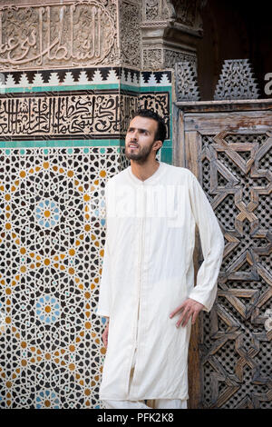 Young muslim man in traditional clothing wearing long white shirt standing in front of the wall with text from Koran and arabesque decoration with flo Stock Photo