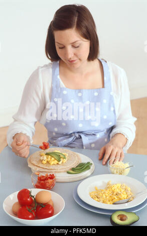Woman spooning tomatoes on tortilla, on a table with bowls of grated cheese, scrambled eggs, tomatoes, boiled eggs, and a halved avocado Stock Photo