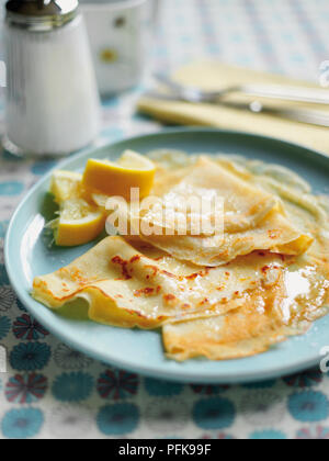 Crepes served with melted butter and lemon slices, on a plate, sugar shaker and cutlery in background Stock Photo