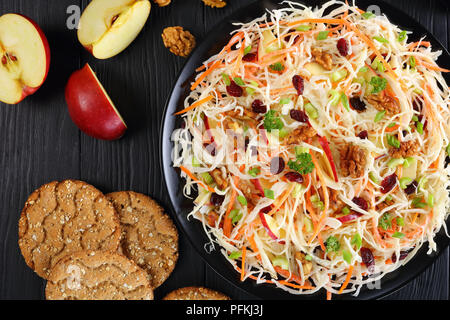 close-up of Apple Cranberry and walnuts Coleslaw salad on a black plate on black wooden table with red apple, multigrain crispbread at background, vie Stock Photo