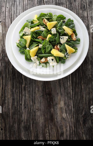 fresh Baby spinach leaves, pineapple slices, blue cheese chunks and walnuts salad on a white plate on dark wooden table, vertical view from above Stock Photo