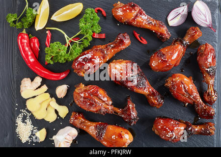 grilled crispy juicy teriyaki chicken drumsticks sprinkled with sesame seeds on black stone tray on wooden table with parsley, garlic and lemon slices Stock Photo