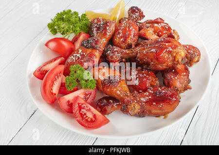 close-up of tasty grilled crispy juicy teriyaki chicken drumsticks sprinkled with sesame seeds on white plate with tomatoes, parsley and lemon slices, Stock Photo