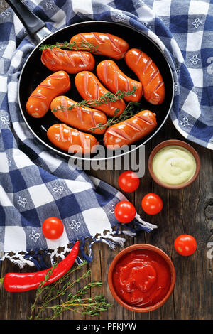 delicious hot fried juicy sausages with fresh thyme on frying pan on dark wooden kitchen table with chili pepper, ketchup and aioli sauce, vertical vi Stock Photo