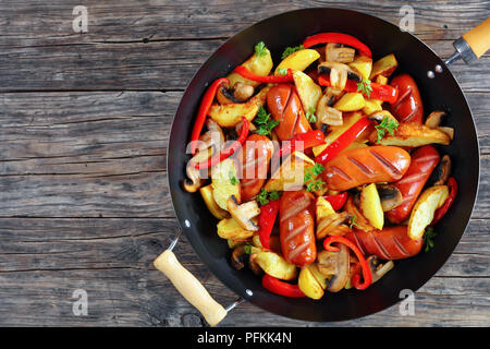 delicious grilled sausages with potato wedges, red bell pepper pieces, mushrooms and parsley in a frying pan, view from above, close-up Stock Photo