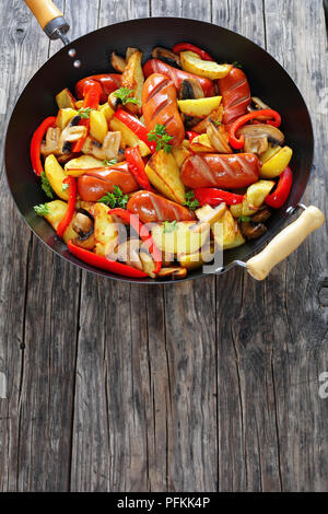 delicious one pot meal - grilled sausages with potato wedges, red bell pepper pieces, mushrooms and parsley in a frying pan, vertical view from above Stock Photo