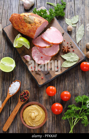 delicious ham cut in slices with spices, parsley and lime on dark wooden cutting board on old wooden table, vertical view from above Stock Photo
