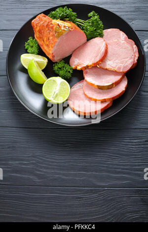 delicious ham cut in slices served with parsley and fresh citrus wedges on black plate on black wooden table, vertical view from above Stock Photo