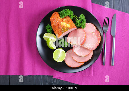 delicious ham cut in slices served with parsley and fresh lime wedges on black plate on  table mat with silver fork and knife, on black wooden table,  Stock Photo