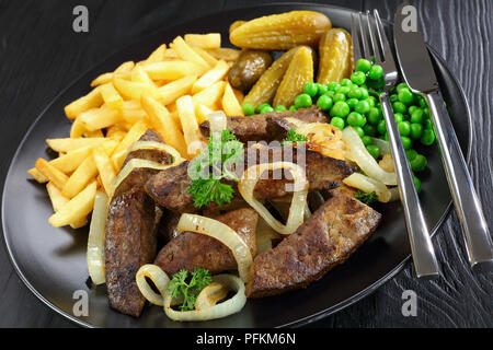 close- up of delicious fried calves liver pieces served with french fries, pickled cucumbers and green peas on black plate with fork and knife, on bla Stock Photo