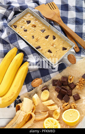 uncooked banana bread with walnuts and chocolate pieces in a loaf pan on wooden table with kitchen towel, spatulas and ingredients at background, vert Stock Photo