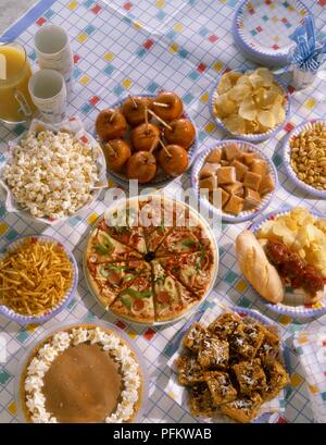 Selection of party food dishes on a table, including pizza slices, cakes, toffee apples, crisps and popcorn on paper plates, orange juice and paper cups Stock Photo