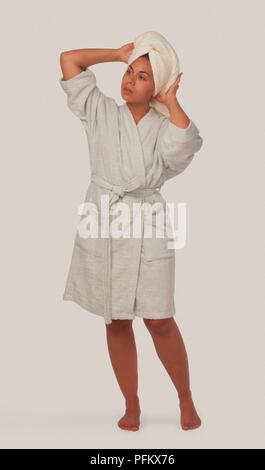 Woman in white bathrobe adjusting towel wrapped around her head, standing, looking away Stock Photo