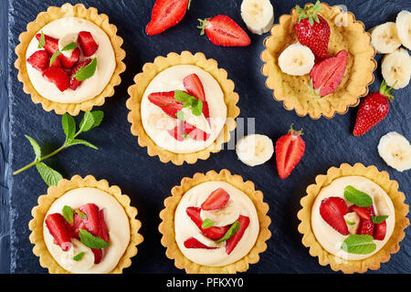 tartlets with strawberries, banana slices loaded with cream diplomat on a black slate plate, view from above, close-up Stock Photo