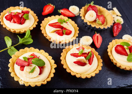 tartlets with strawberries, banana slices loaded with cream diplomat on a black slate plate, view from above, close-up Stock Photo